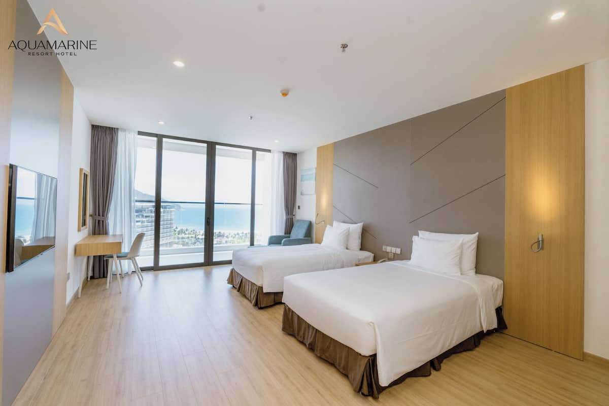 A hotel room with two beds, a desk, and a large window offering a view of the sea.