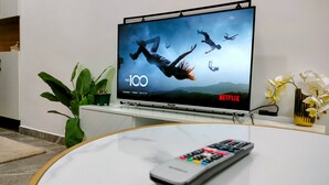 Un televisor de pantalla plana sobre un soporte mostrando 'The 100' en Netflix, con un control remoto sobre una mesa de vidrio al frente.