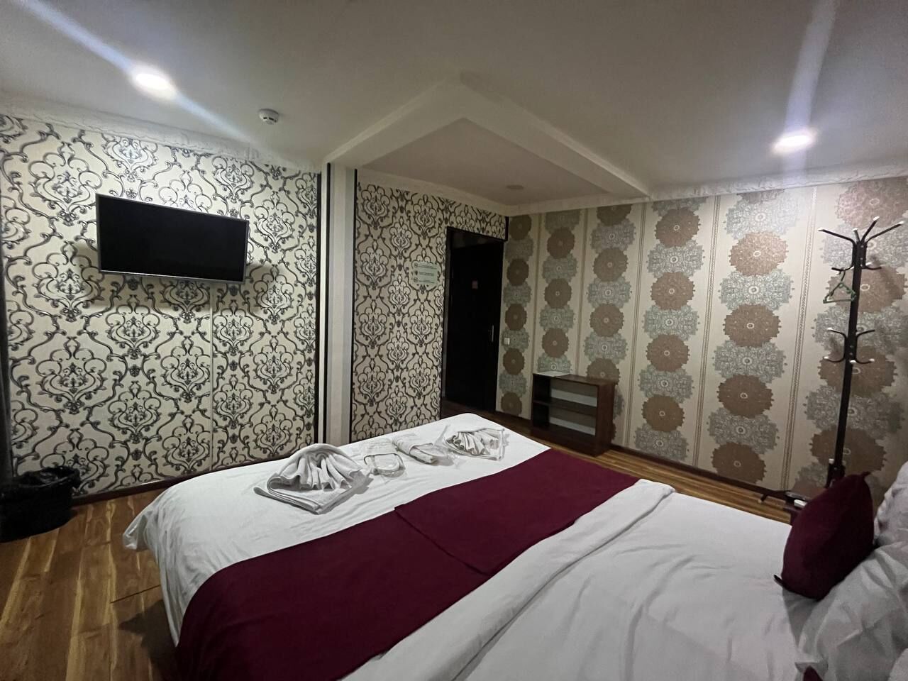 A hotel room with a large bed, a flat-screen TV mounted on the wall, patterned wallpaper, and a floor lamp.