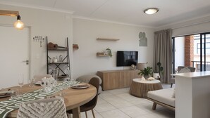 A modern living room with a dining area, a TV, and a large window with curtains. - Ballito Hills Unit 79 (Ballito)