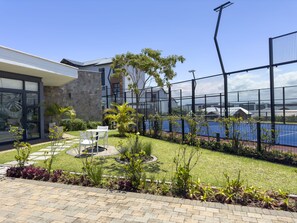 A modern building with a stone facade, a glass entrance, and a well-maintained garden with a tennis court.