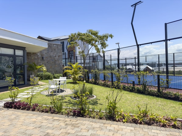 A modern building with a stone facade, a glass entrance, and a well-maintained garden with a tennis court.