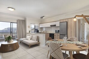 A modern living room with a sofa, a dining table set for four, and a kitchen area with a refrigerator and stove. - Ballito Hills Unit 79 (Ballito)