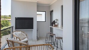 A balcony with a wicker chair, a small table, and a TV mounted on the wall. - Ballito Hills Unit 79 (Ballito)