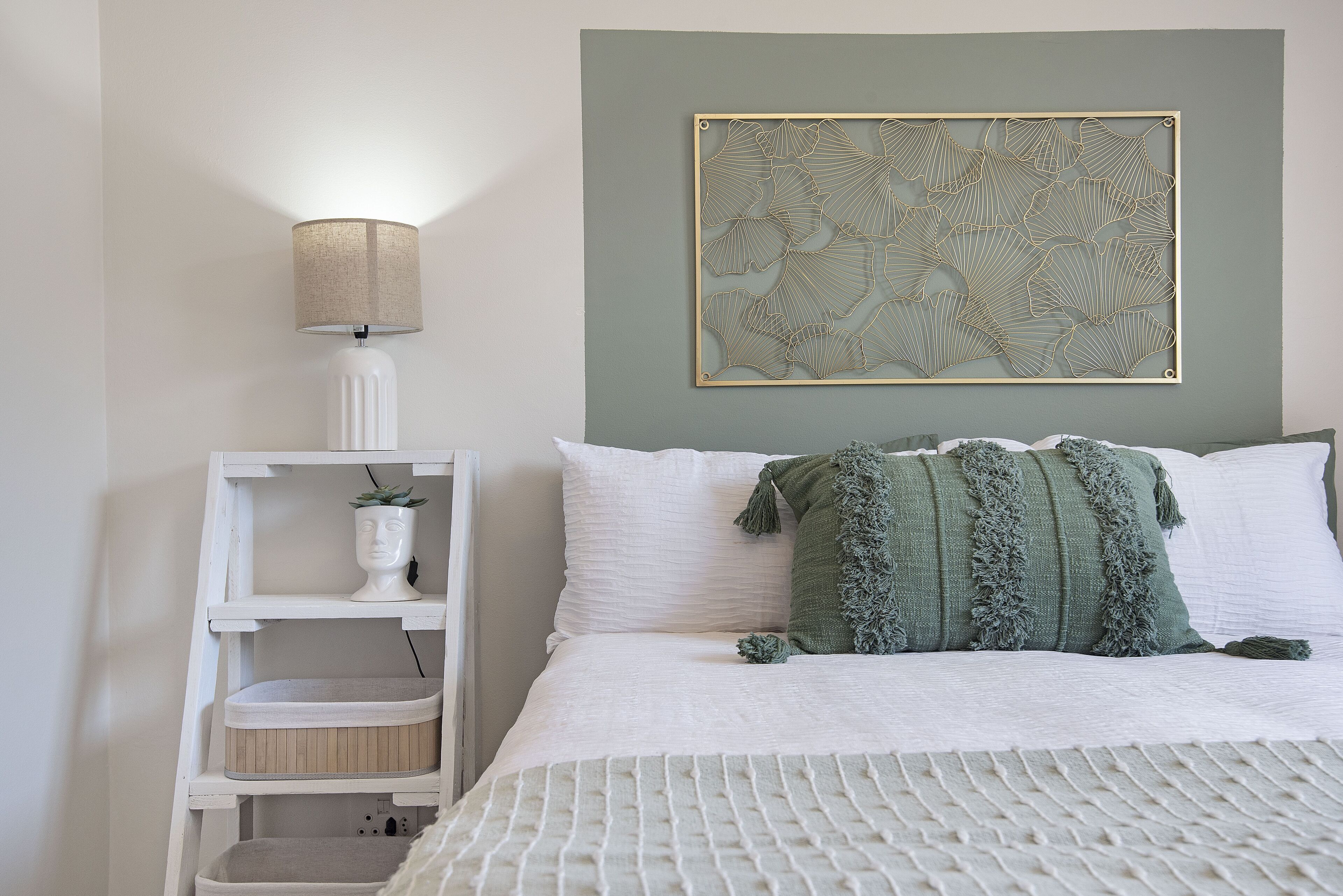 A neatly arranged bedroom with a bed, a bedside lamp, a wall art piece, and a shelf with a plant.