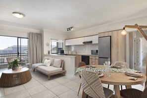 A modern living room with a sofa, a dining table set for four, and a kitchen area with a refrigerator and stove. - Ballito Hills Unit 79 (Ballito)