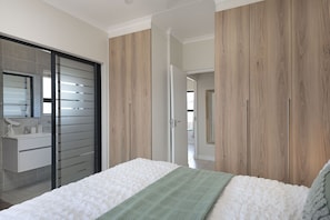 A bedroom with a bed, a green throw, wooden wardrobe, and a bathroom with a mirror and sink.