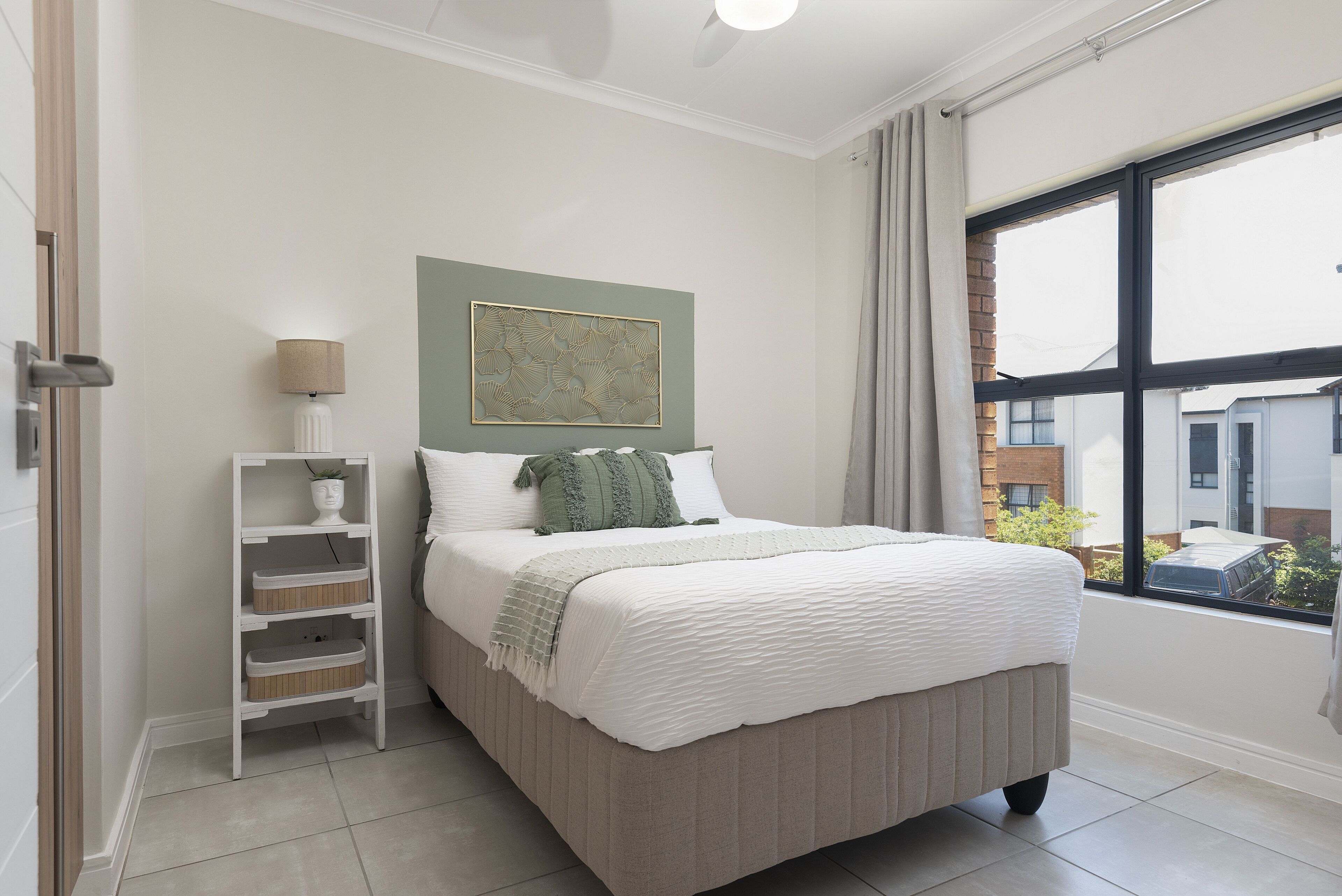 A neatly arranged bedroom with a bed, a side table with a lamp, a wall-mounted shelf, and a window with a view of buildings.