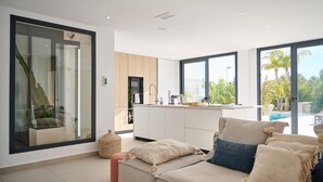 A modern living room with a large window, a sofa, and a kitchen area visible through the glass door. - Casa Palmera - Polop, Costa Blanca (Polop)