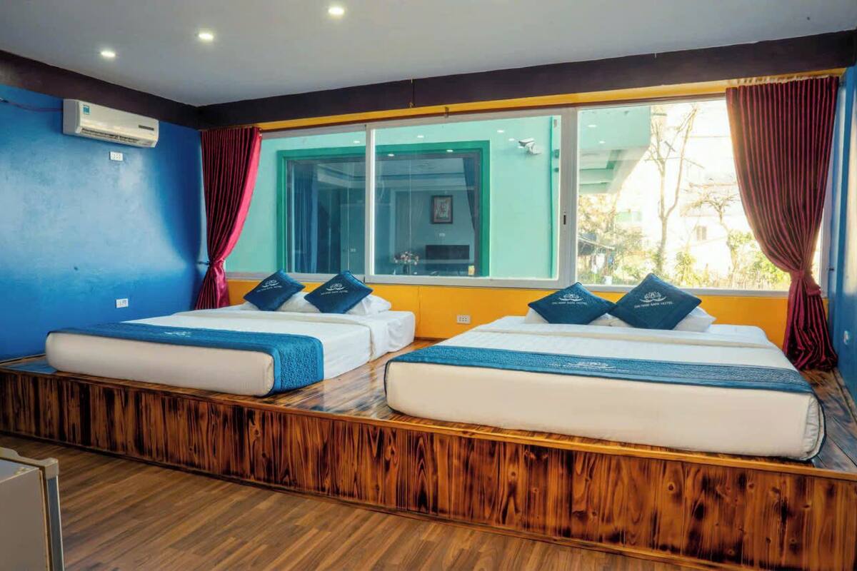 A room with two beds, a wooden platform, a window with curtains, and an air conditioning unit.