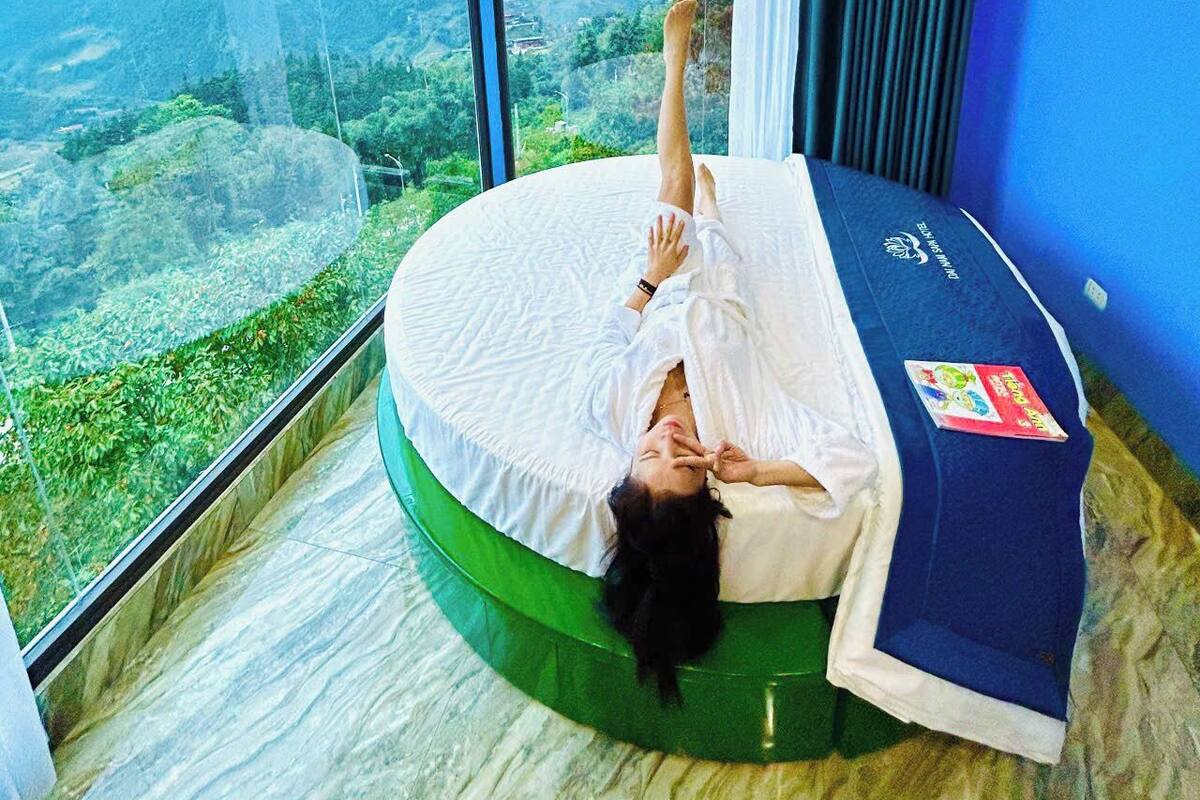 A person lying on a circular bed with a view of a mountainous landscape through large windows.