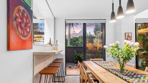 A modern kitchen with a wooden bar and stools, a dining table with a colorful tile runner, and a large window with a view of the outdoors.