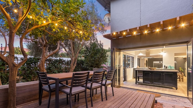 An outdoor deck with a wooden table and chairs, a pergola with string lights, and a view into a modern kitchen through glass doors.