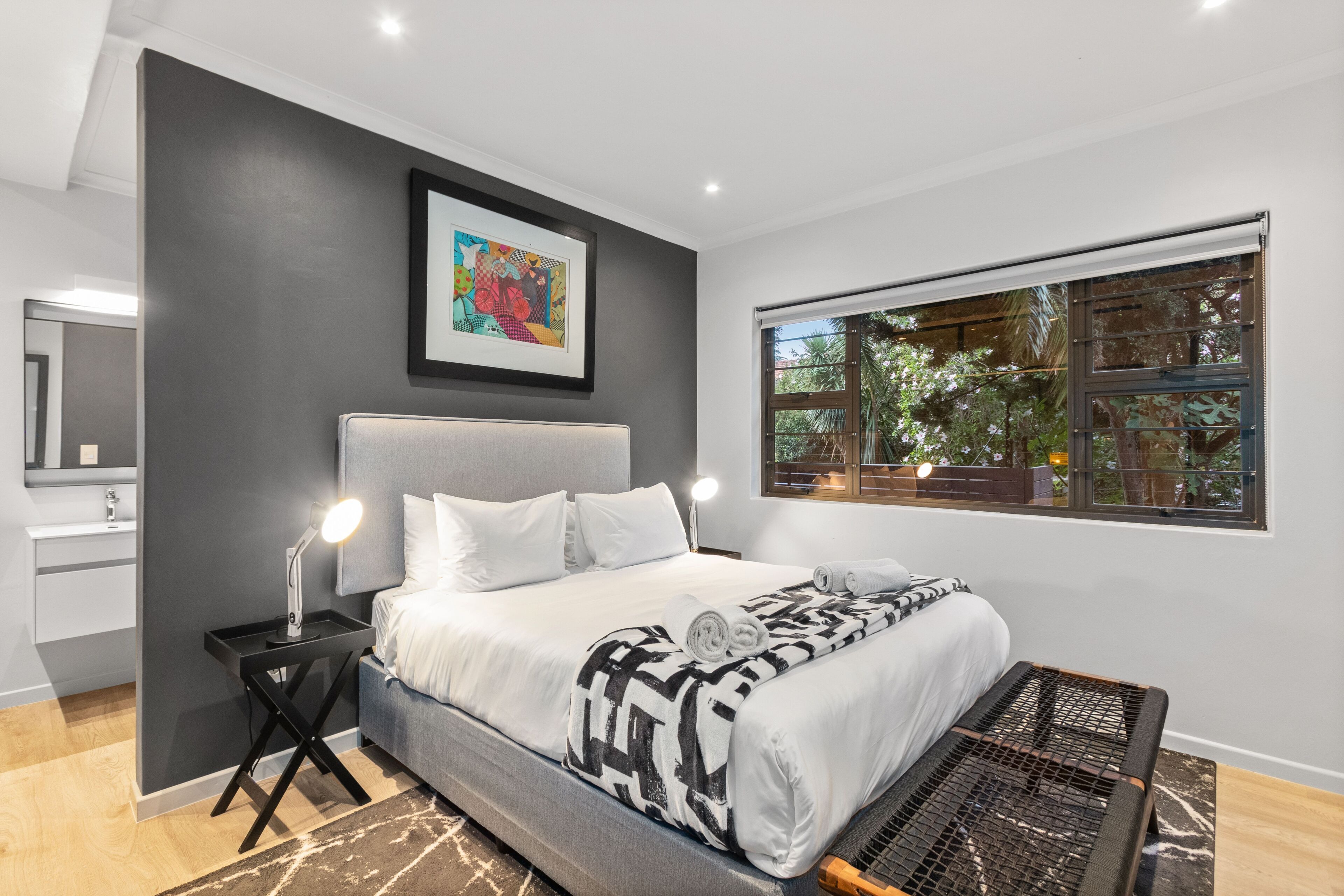 A modern bedroom with a large bed, bedside tables, a nightstand, a window with a view of trees, and a framed artwork on the wall.