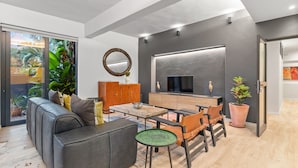 A modern living room with a sofa, coffee table, and chairs. There is a television on the wall and a large window with a view of greenery.
