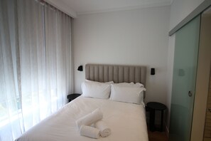 A neatly made bed with white linens and two pillows, a bedside table with a lamp, and a glass door to the right. - The Barracks 1 Bedroom (Cape Town)