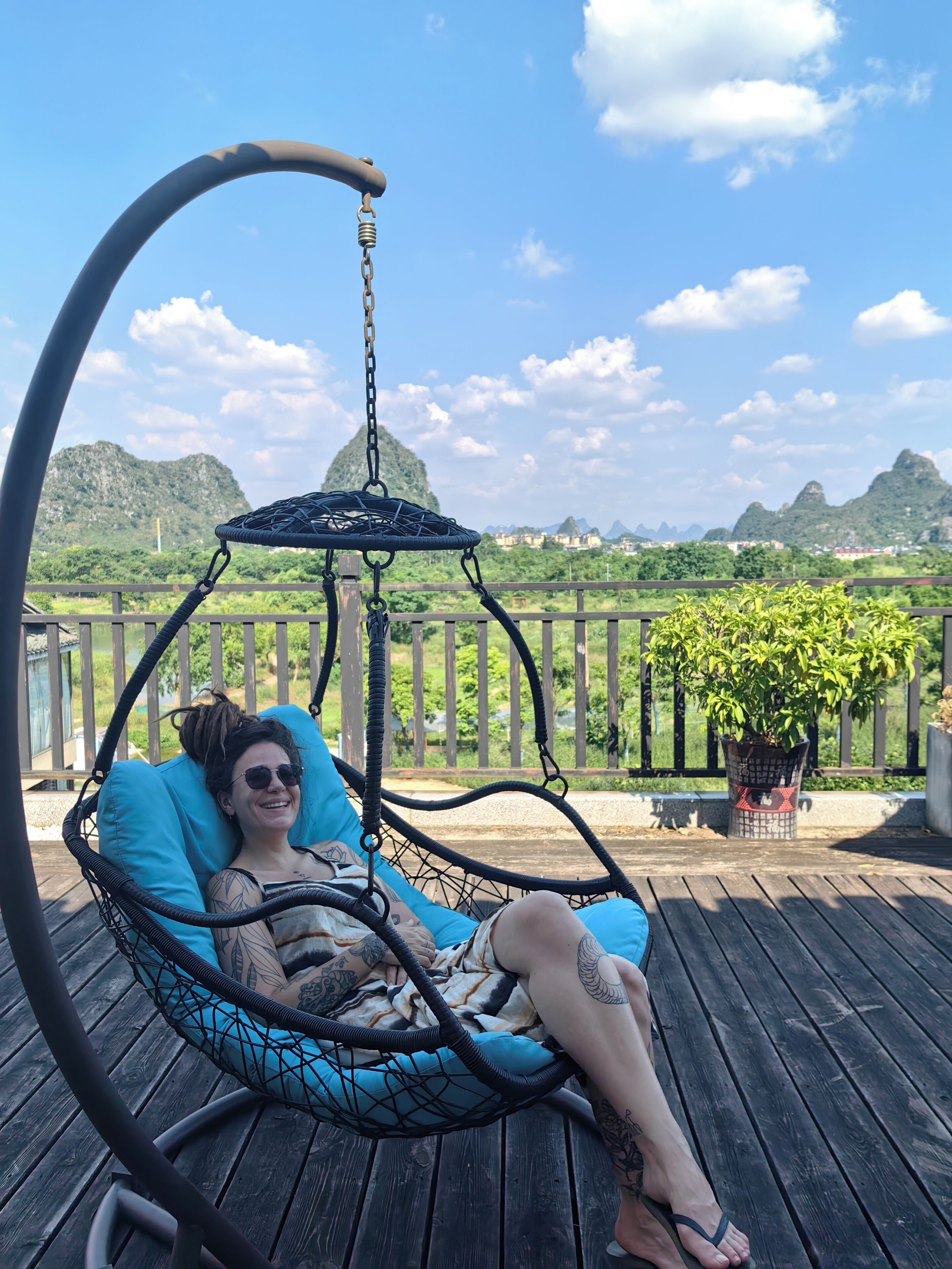 A person relaxing in a hanging chair on a wooden deck with a view of mountains and greenery.