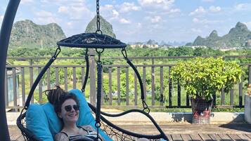 A person relaxing in a hanging chair on a wooden deck with a view of mountains and greenery.