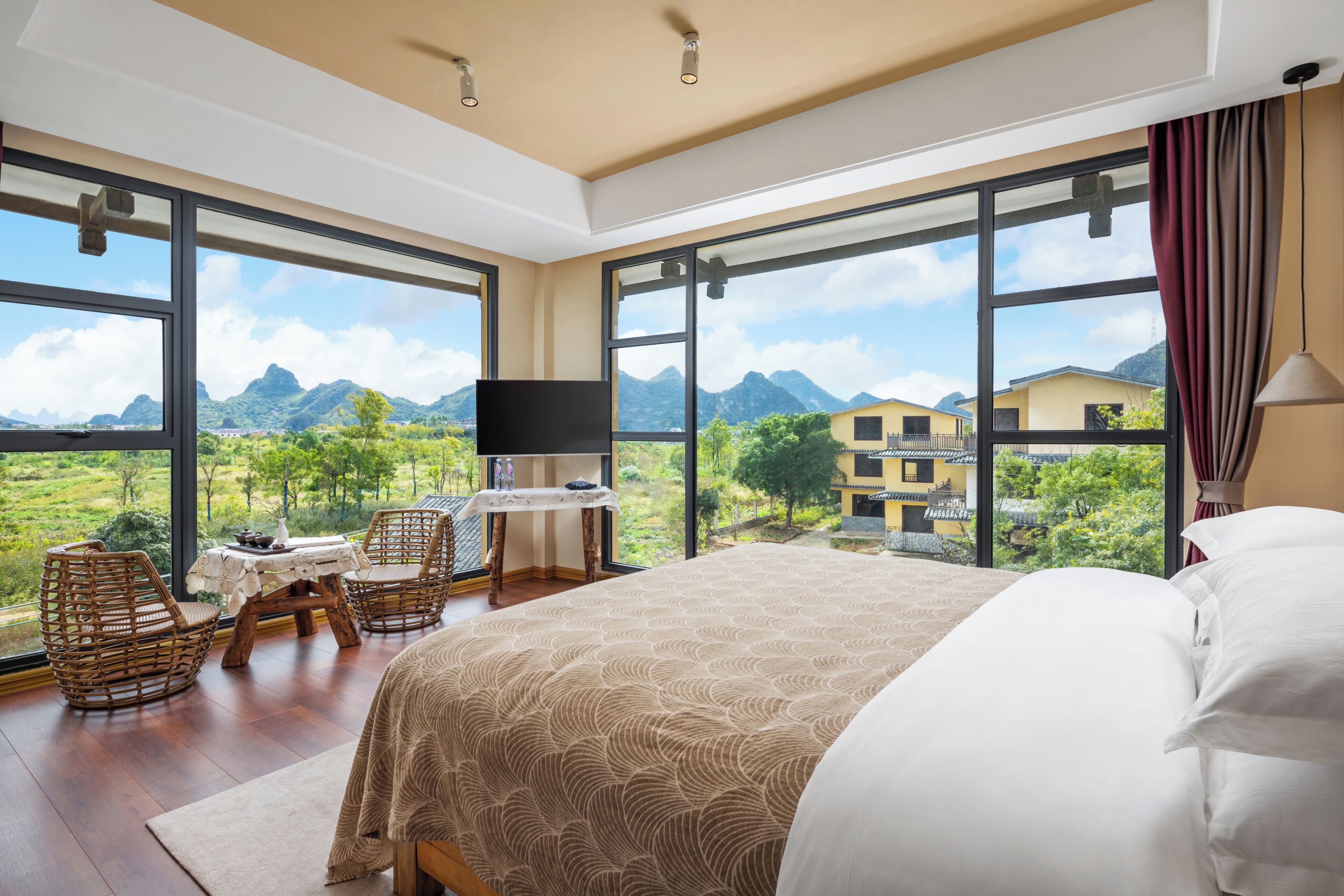 A bedroom with a bed, a desk, and chairs, large windows with a mountain view, and a television.