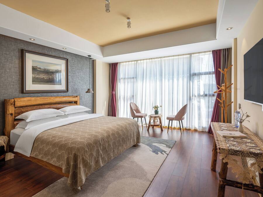 A bedroom with a bed, bedside table, chairs, a table, a TV, and a window with curtains.