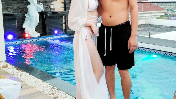 Una pareja de pie junto a una piscina, con un muro de piedra y una estatua al fondo.