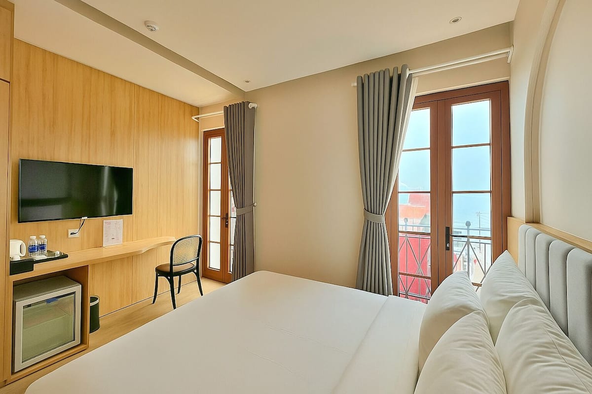 A hotel room with a large bed, a TV mounted on the wall, a desk with a chair, and a window with curtains.