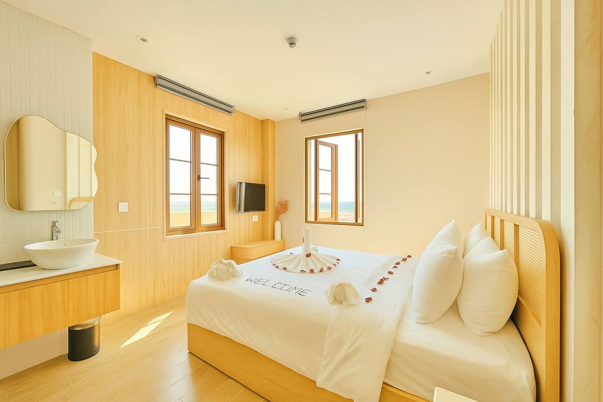 A modern bedroom with a wooden floor, a white bed with a 'WELCOME' sign, a wooden headboard, a large mirror, and a view of the beach through the windows.