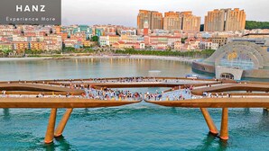 A waterfront promenade with people walking, a modern bridge structure, and a cityscape with colorful buildings in the background. - HAN Lagoon Sunset Hotel (Phu Quoc)