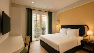 Habitación de hotel con cama, escritorio, silla, televisor y balcón con cortinas.