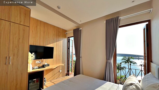 A modern hotel room with a large bed, a TV on a wooden cabinet, and a balcony with a view of the sea. - HAN Lagoon Sunset Hotel (Phu Quoc)