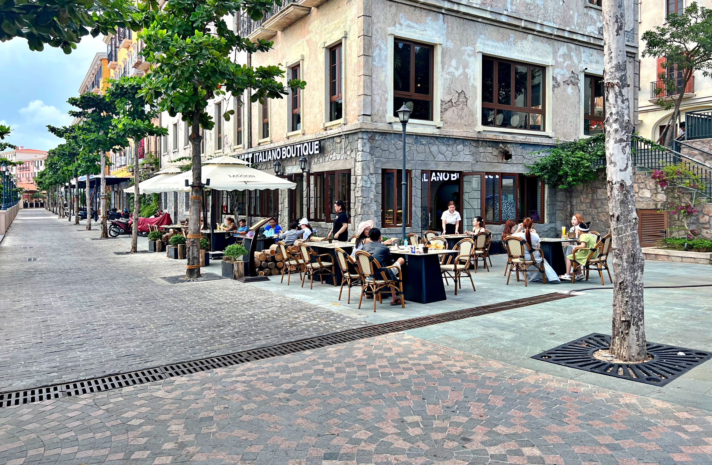 Un café en plein air avec des tables et des chaises, un bâtiment en pierre sur lequel on peut lire l’inscription « ITALIANO BOUTIQUE », et un arbre au premier plan.