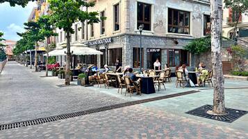 Un café en plein air avec des tables et des chaises, un bâtiment en pierre sur lequel on peut lire l’inscription « ITALIANO BOUTIQUE », et un arbre au premier plan.