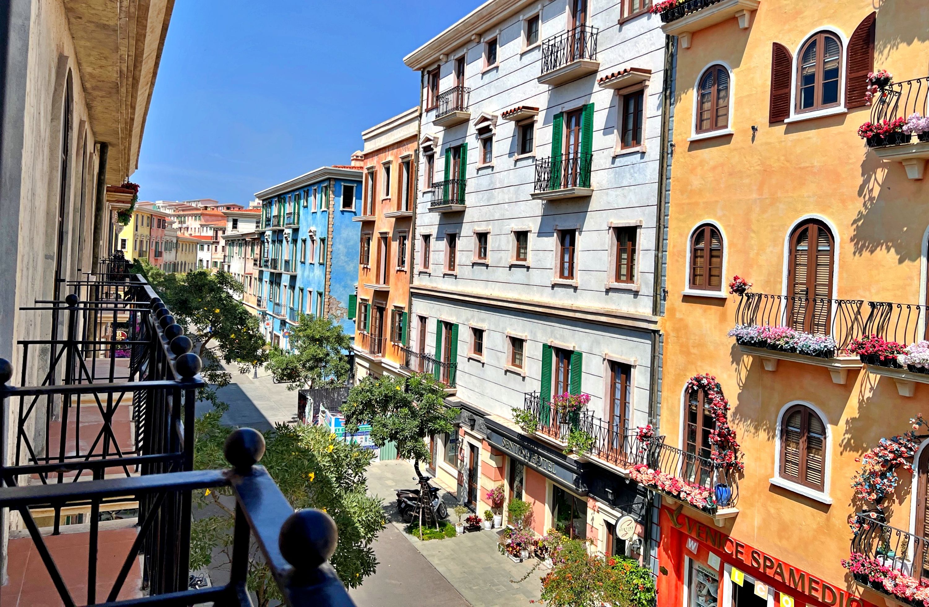 Uma fileira de edifícios coloridos com varandas, céu claro e uma placa com a inscrição "VENICE SPAMEDIO".