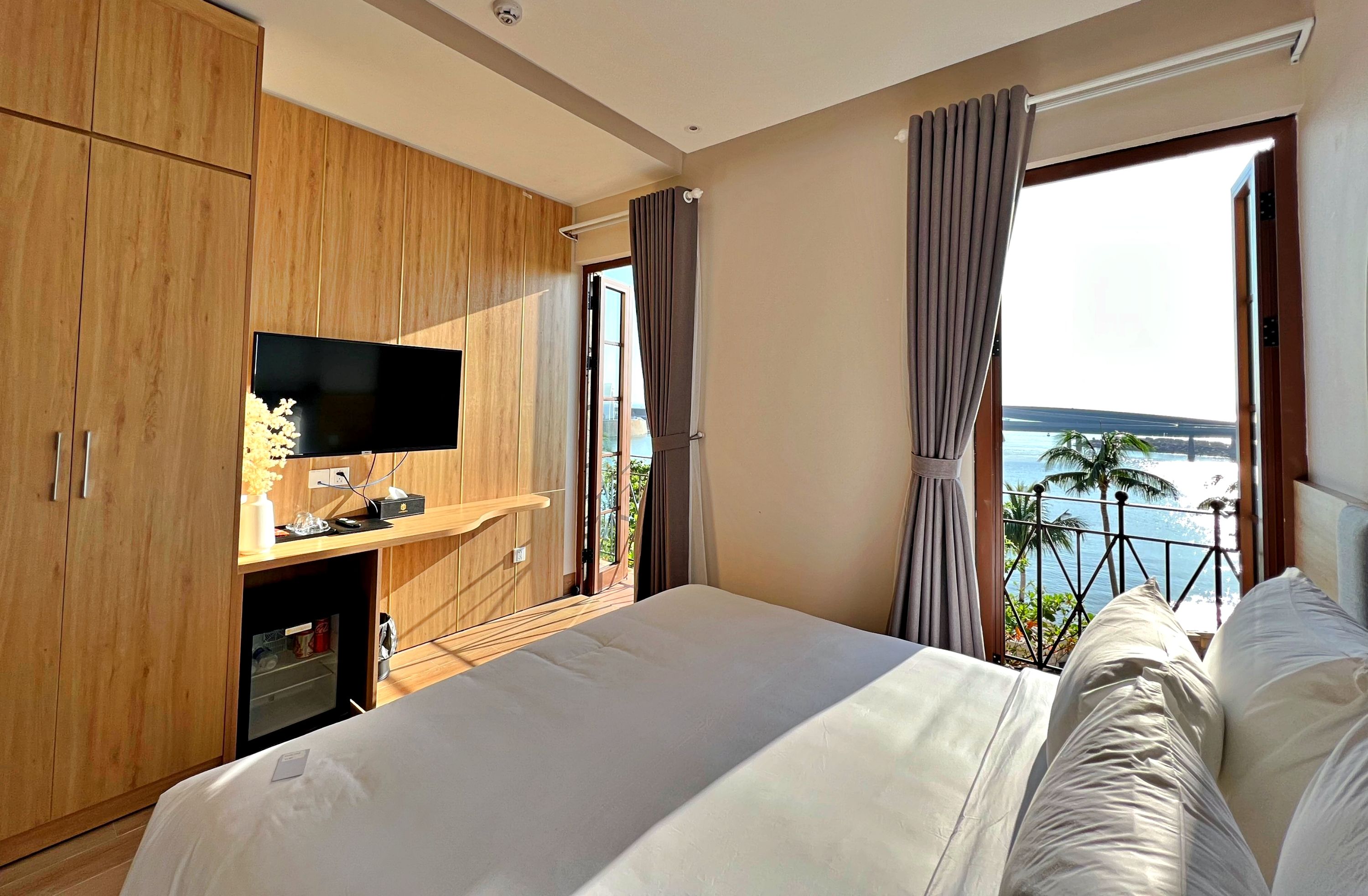 Ein modernes Hotelzimmer mit einem großen Bett, einem Fernseher auf einer Holzkassette und einem Balkon mit Meerblick.