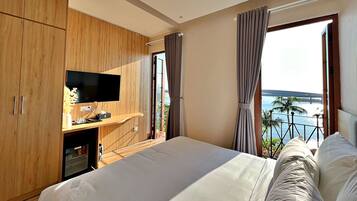 Une chambre d’hôtel moderne avec un grand lit, un téléviseur posé sur une commode en bois et un balcon offrant une vue sur la mer.