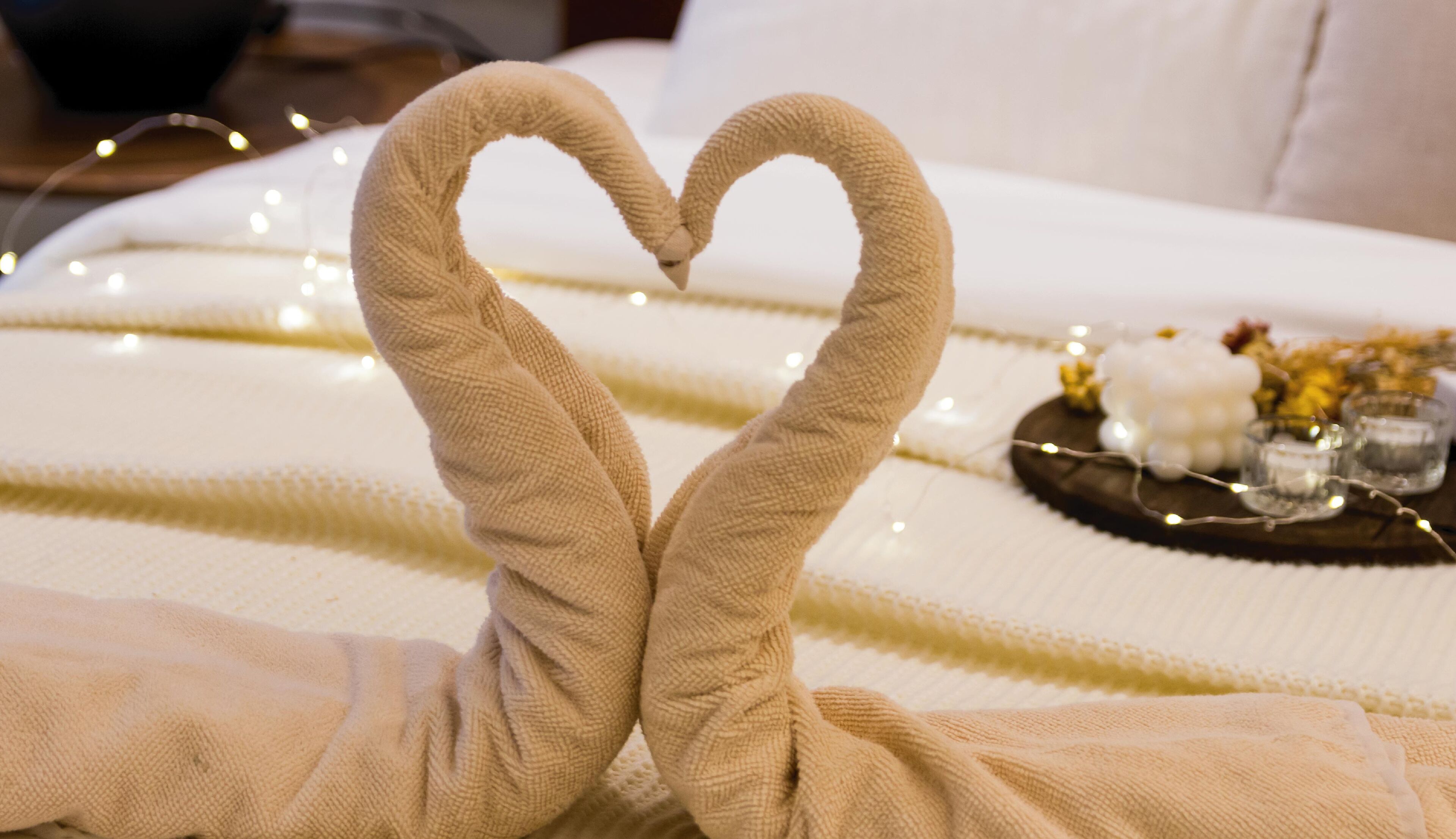 A bed with a heart-shaped towel arrangement and a tray with flowers and candles.