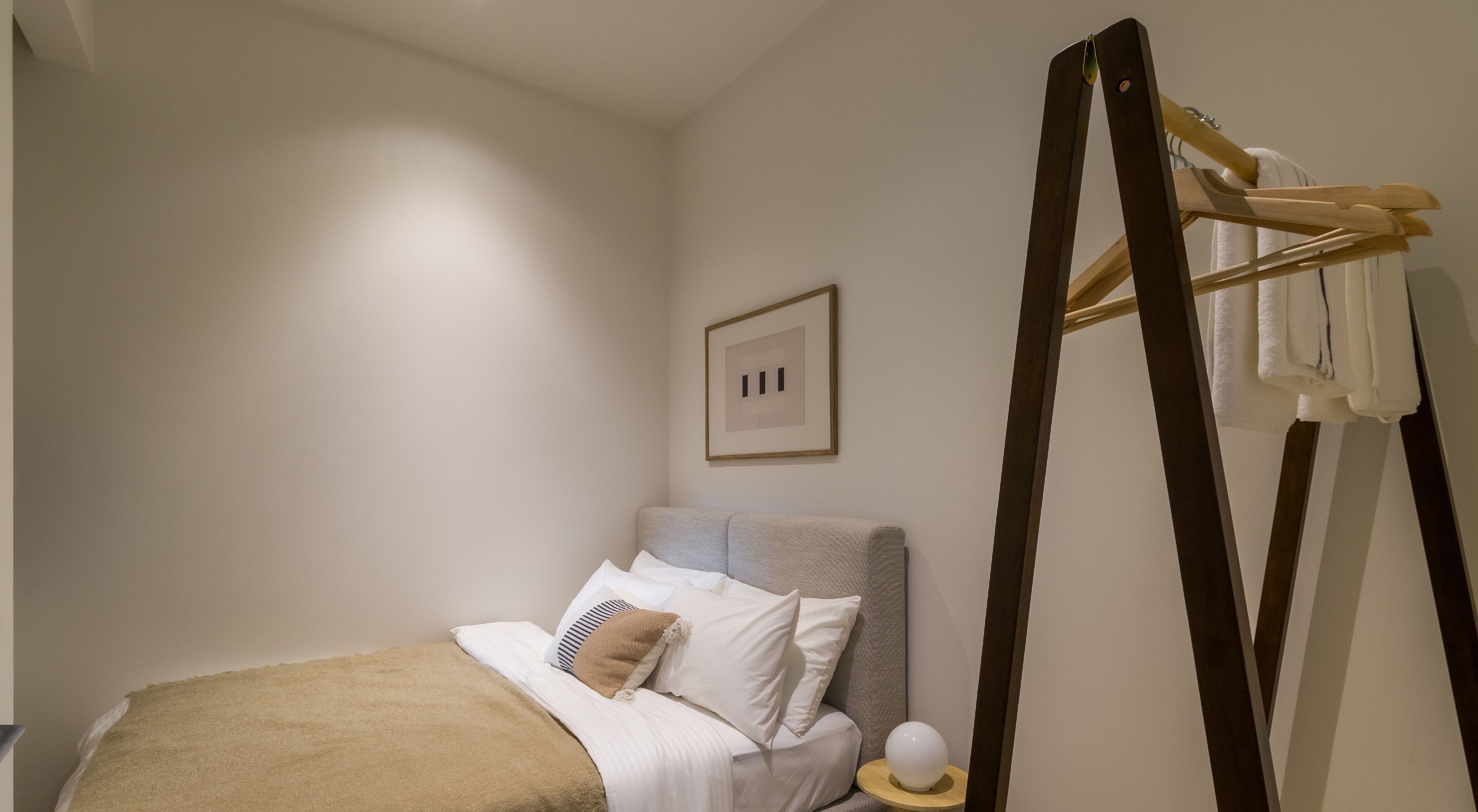 Un dormitorio con cama, almohadas, un perchero, un cuadro enmarcado y una lámpara de noche.