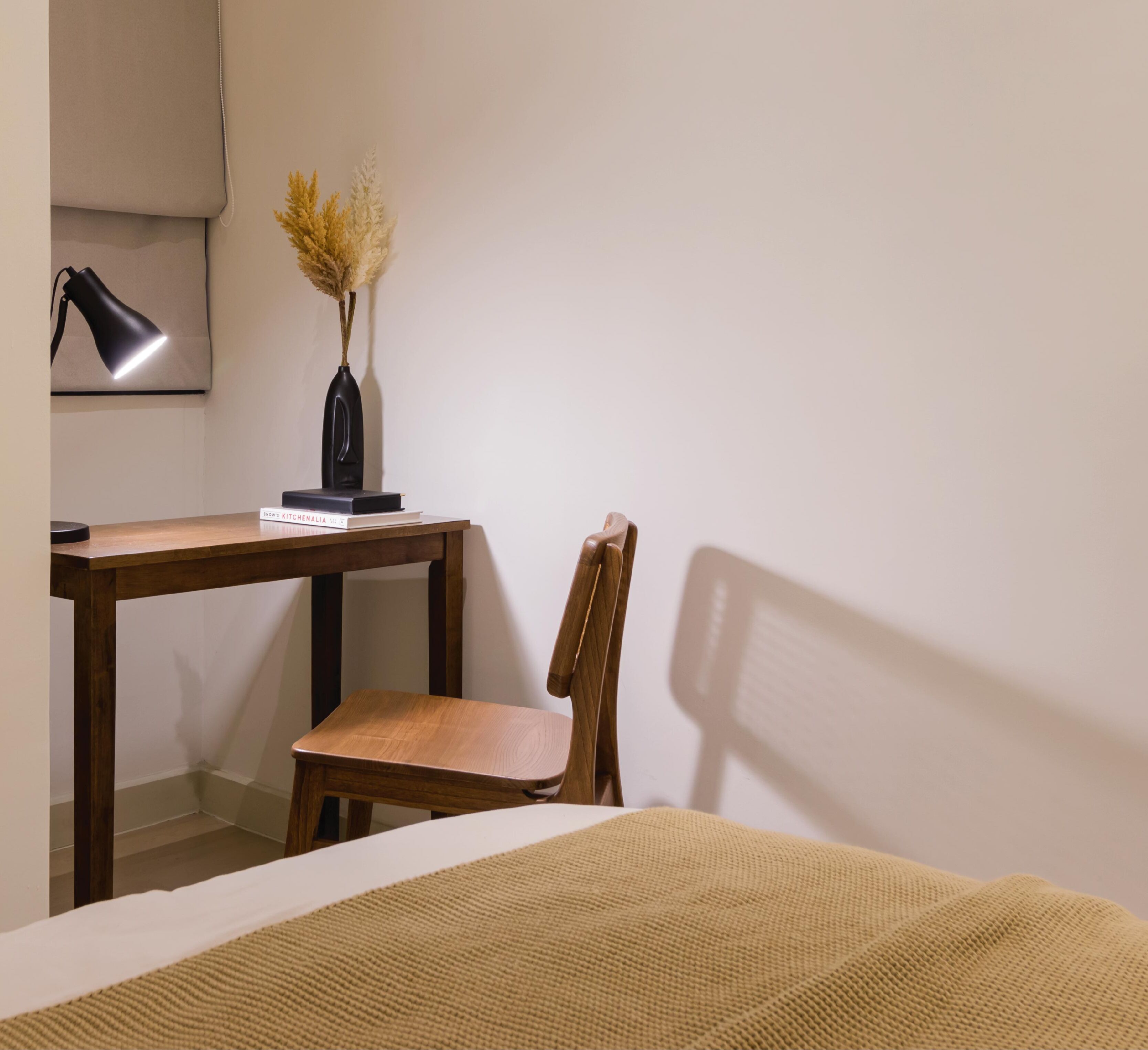 A corner bedroom with a wooden desk, a chair, a bedside table, a lamp, and a vase with dried flowers.