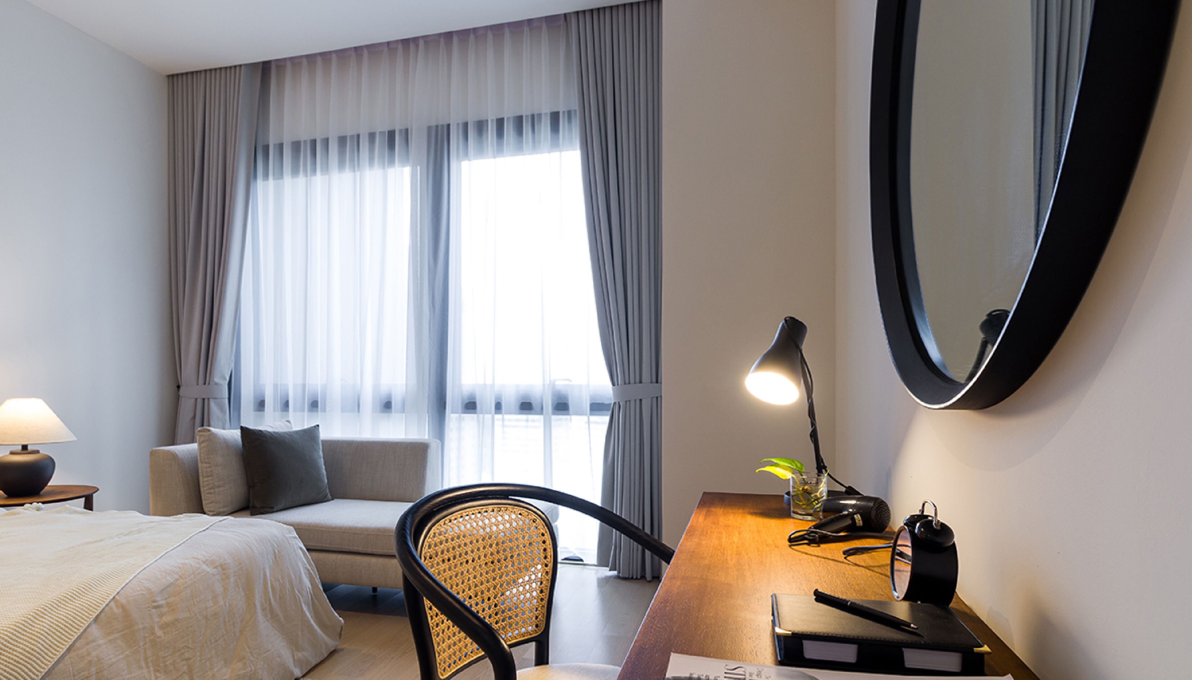 A hotel room with a bed, a desk with a lamp, a chair, a mirror, and a window with curtains.