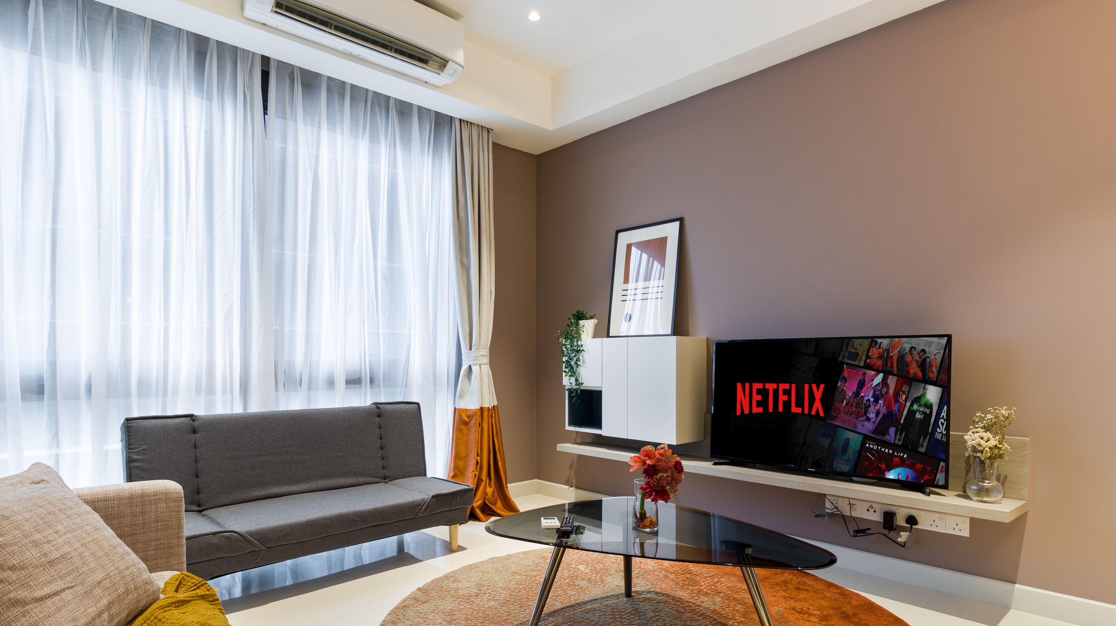 Ein modernes Wohnzimmer mit grauem Sofa, einem Glastisch, einem Flachbildfernseher und einem an der Wand befestigten Regal mit einer Pflanze.