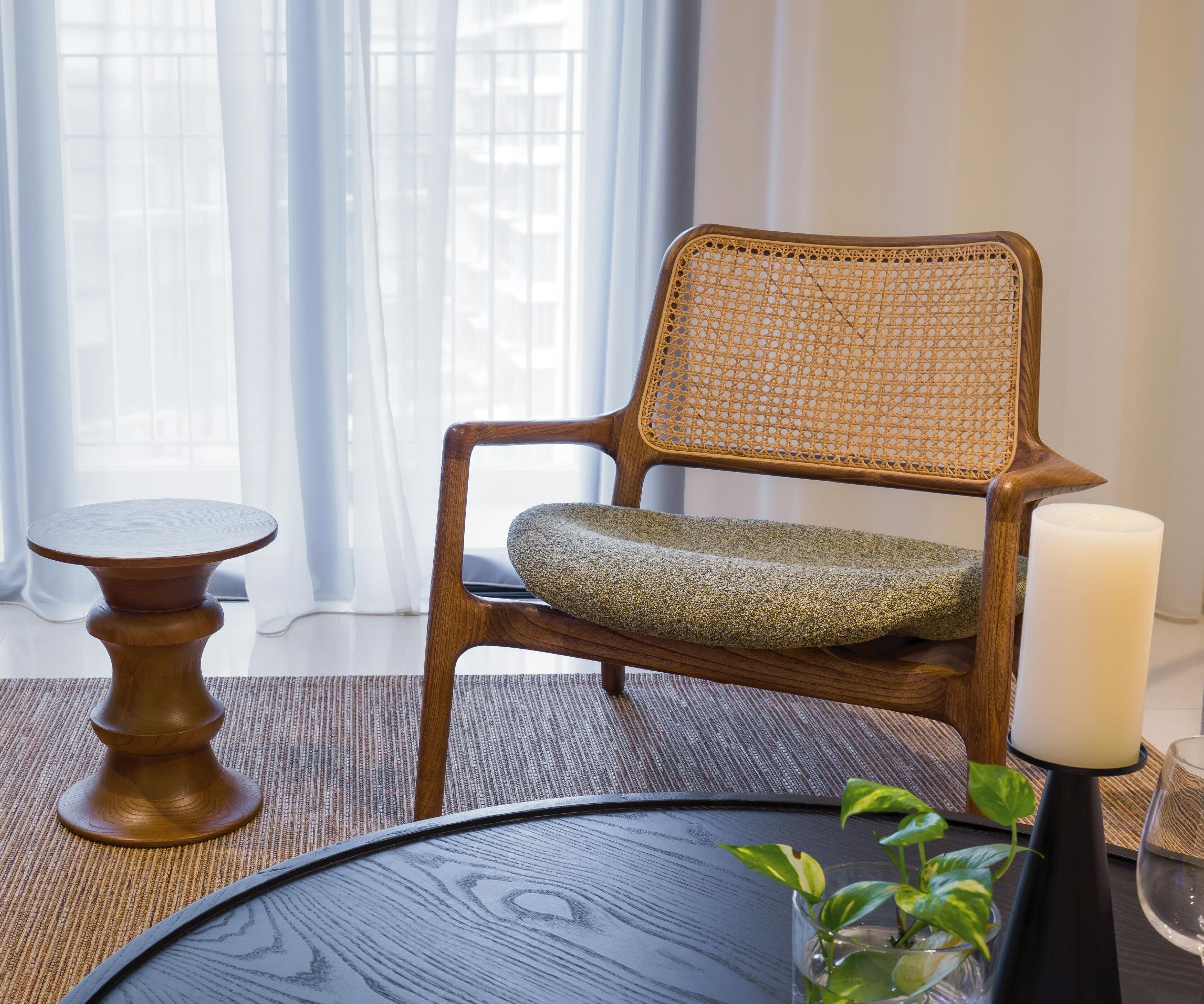 Una silla de madera con respaldo trenzado y asiento acolchado, una mesita redonda pequeña, una vela y un florero de vidrio con una planta.