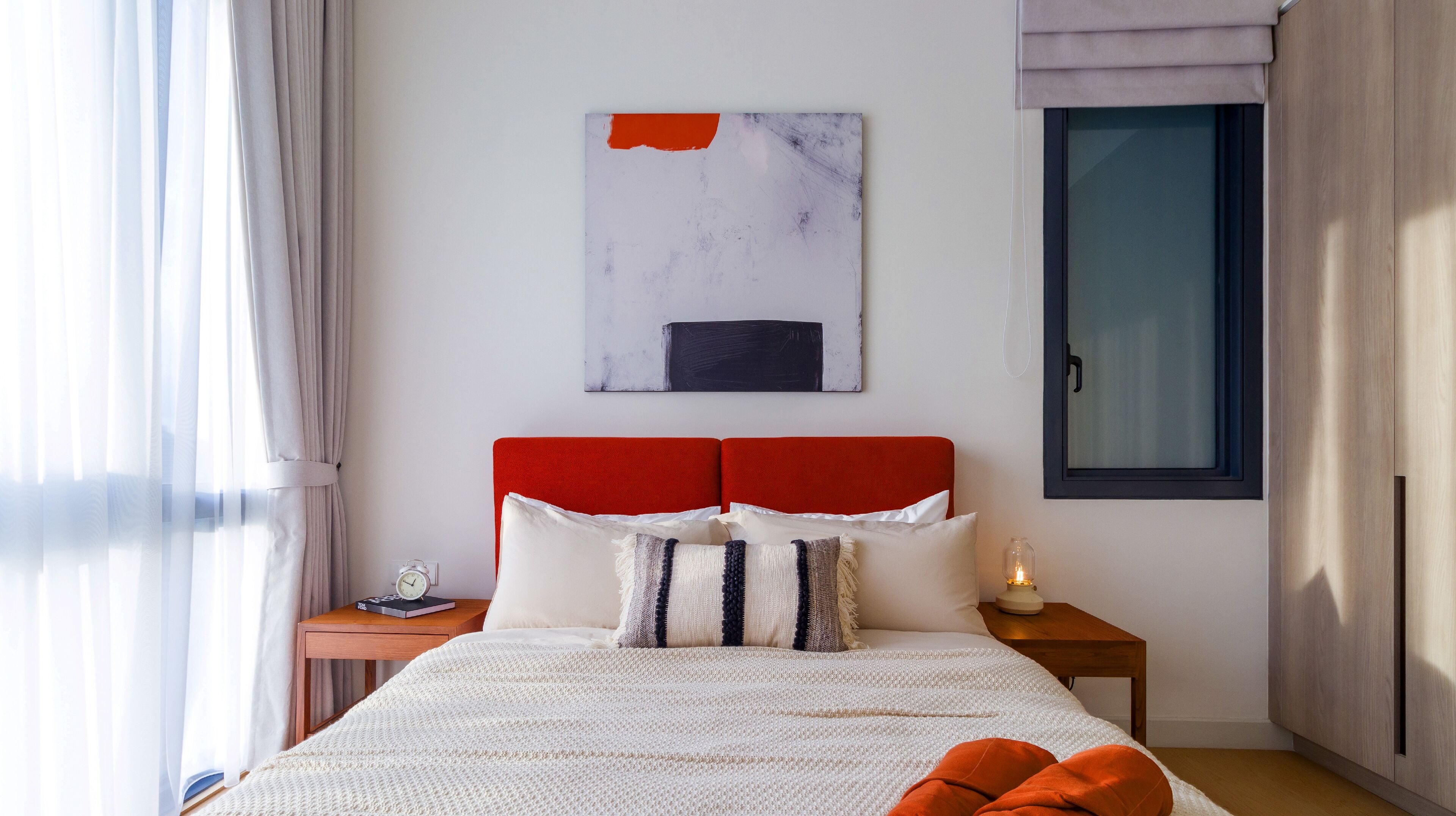 Een moderne slaapkamer met een rode hoofdbord, een groot abstract schilderij, een nachtkastje met een lamp en een raam met gordijnen.