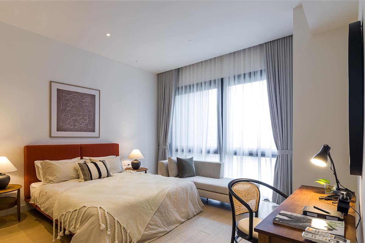 Habitació d'hotel amb llit, sofà, escriptori amb làmpada i finestra gran amb cortines.