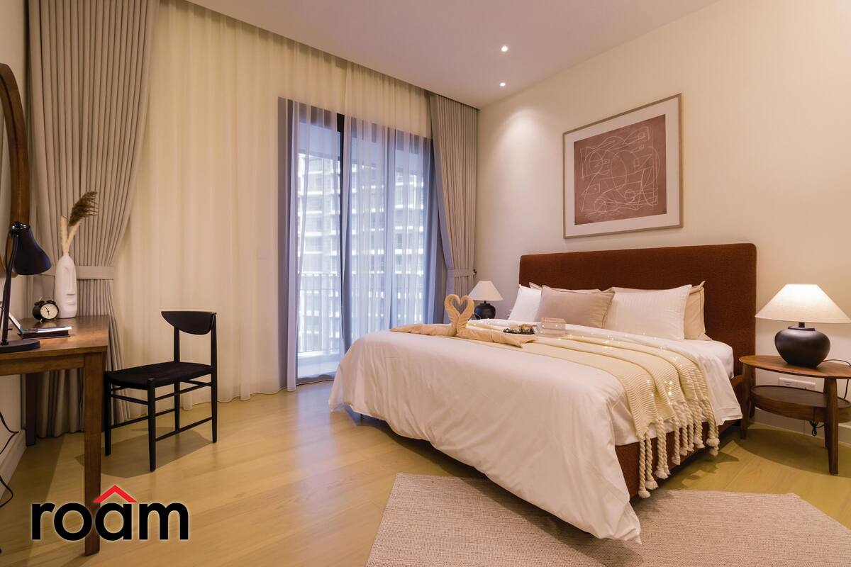 Habitació d'hotel amb llit, escriptori i cadira, una làmpada i finestra amb cortines.