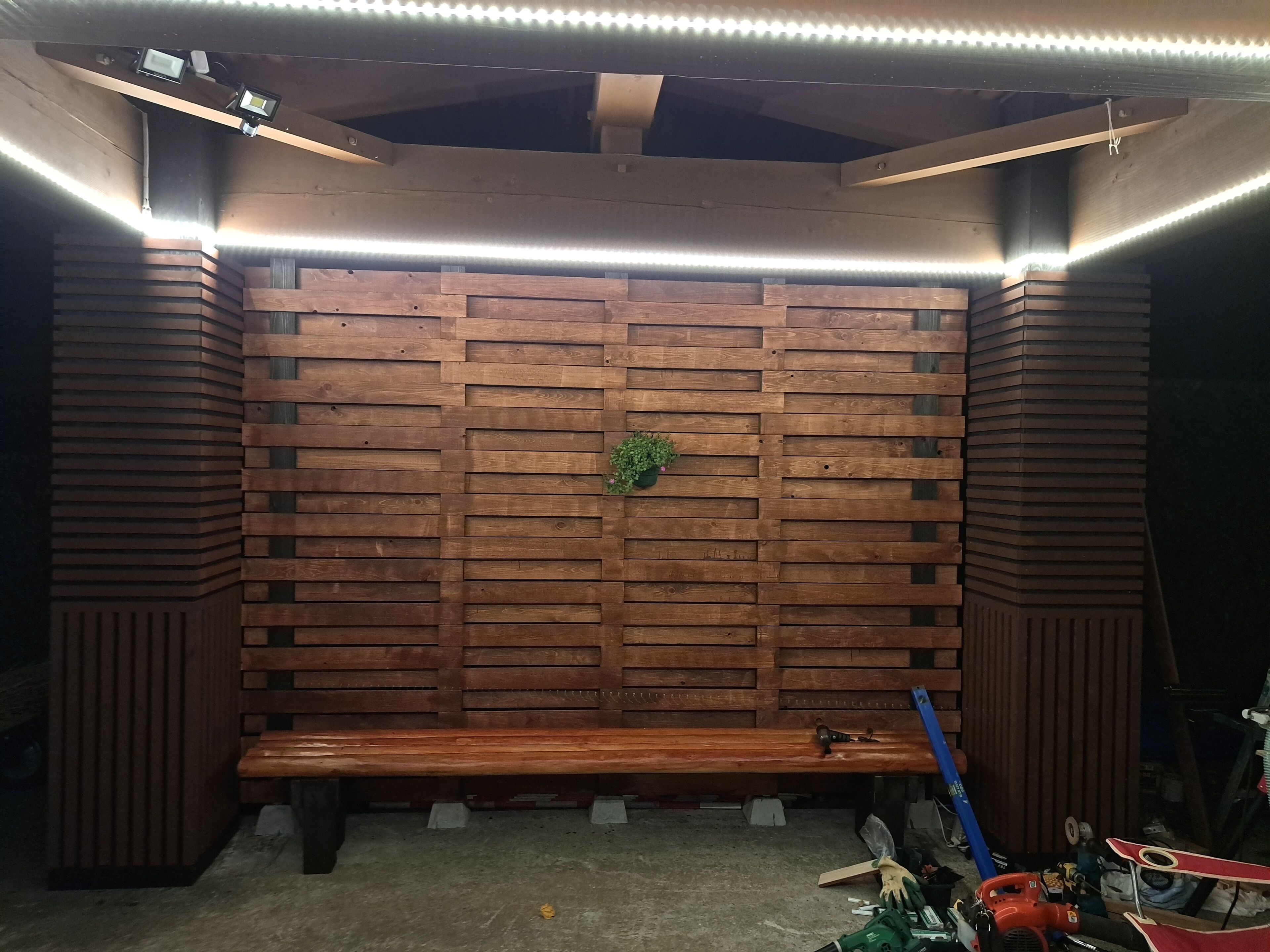 A wooden bench with a green plant in a corner, under a ceiling with exposed beams and LED lighting.