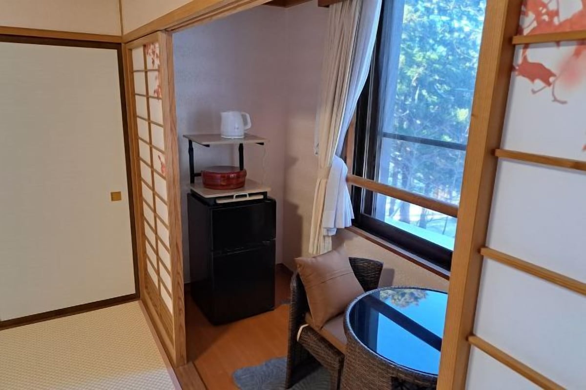 Um quarto japonês tradicional com piso de tatame, uma mesa baixa com almofadas, um móvel preto e uma janela com vista para árvores.