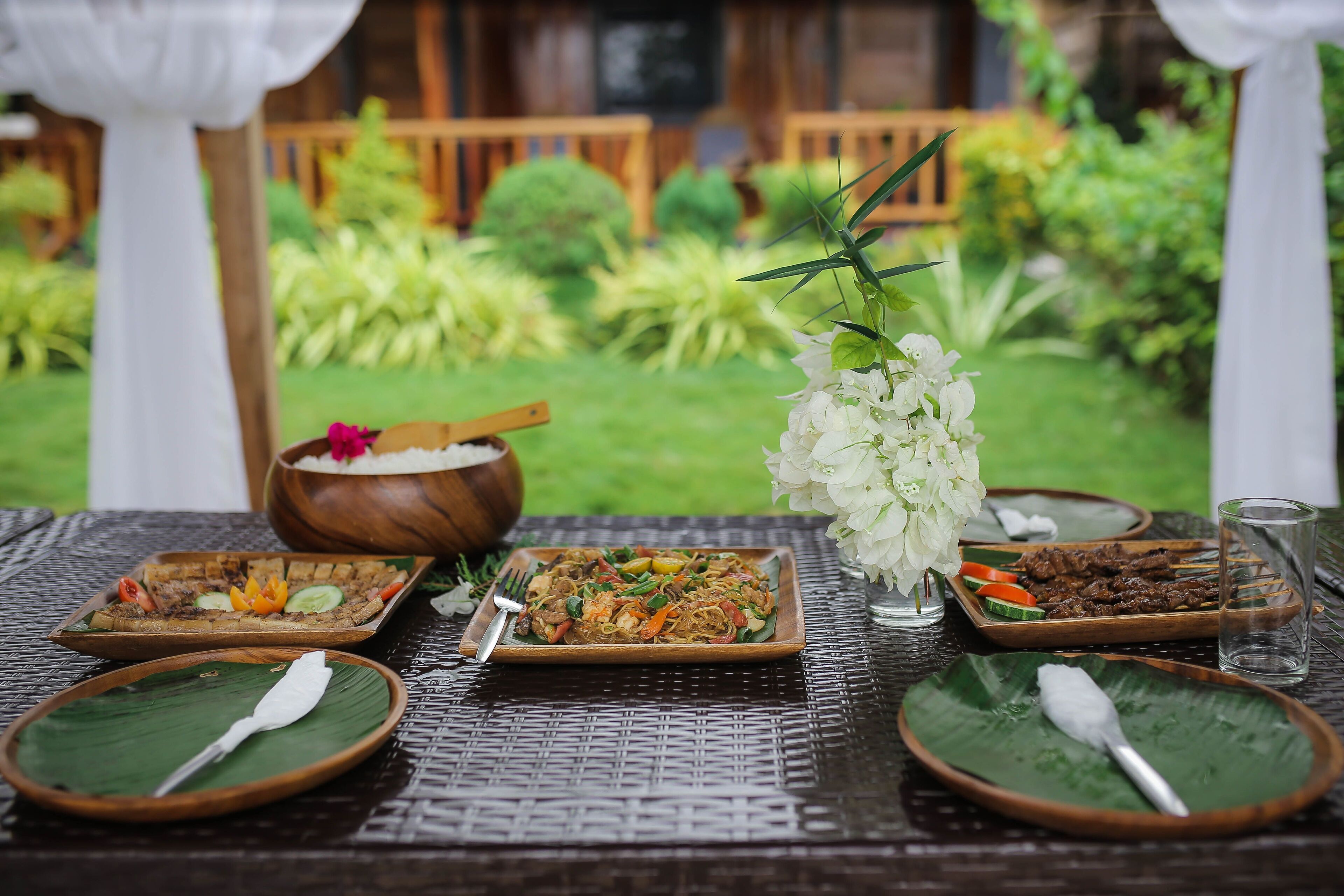 A table set with various dishes, a bowl, and a glass of water, with a garden view in the background.