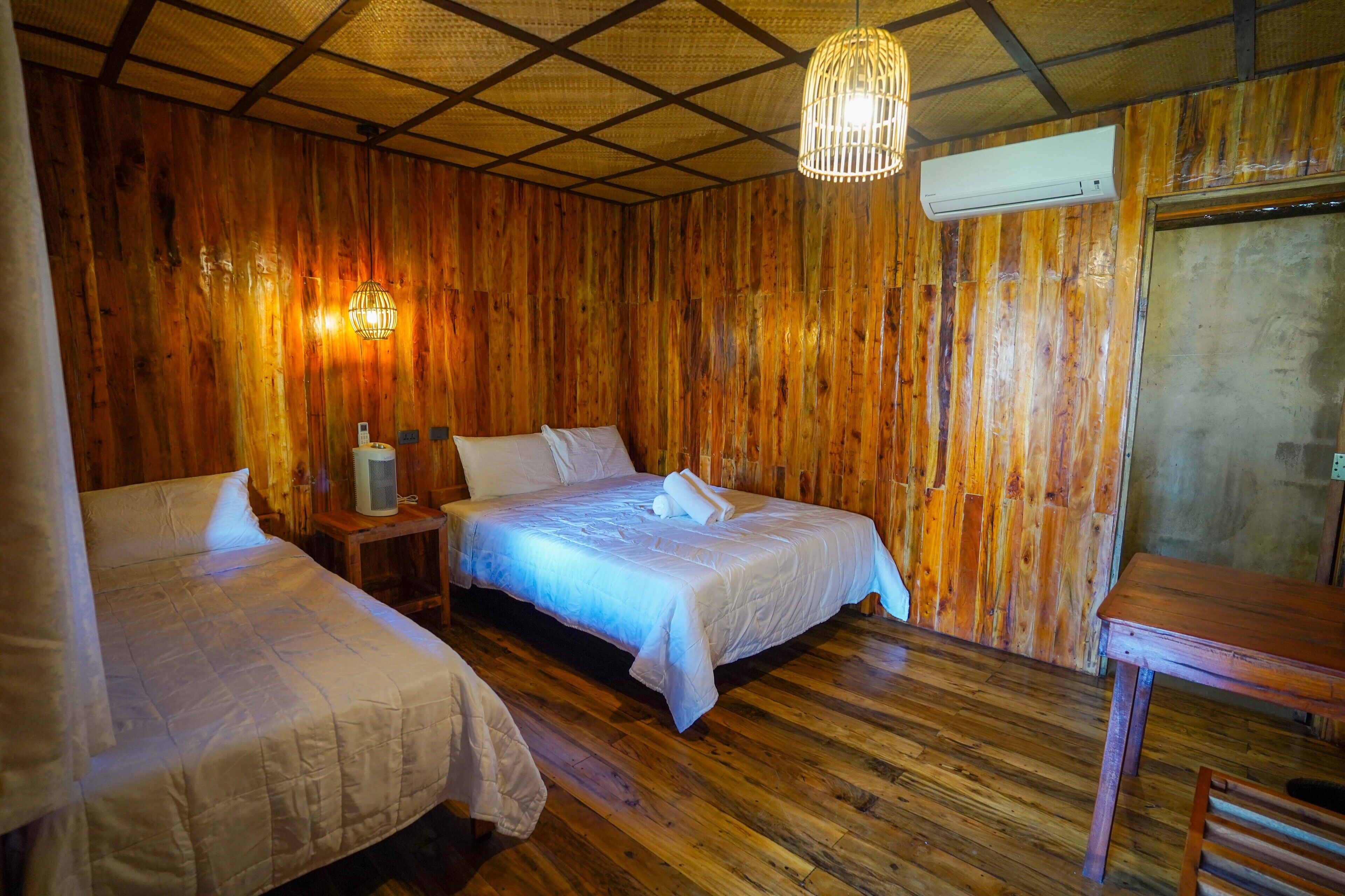 A room with wooden walls and floors, two beds with white linens, a hanging light fixture, and an air conditioning unit.