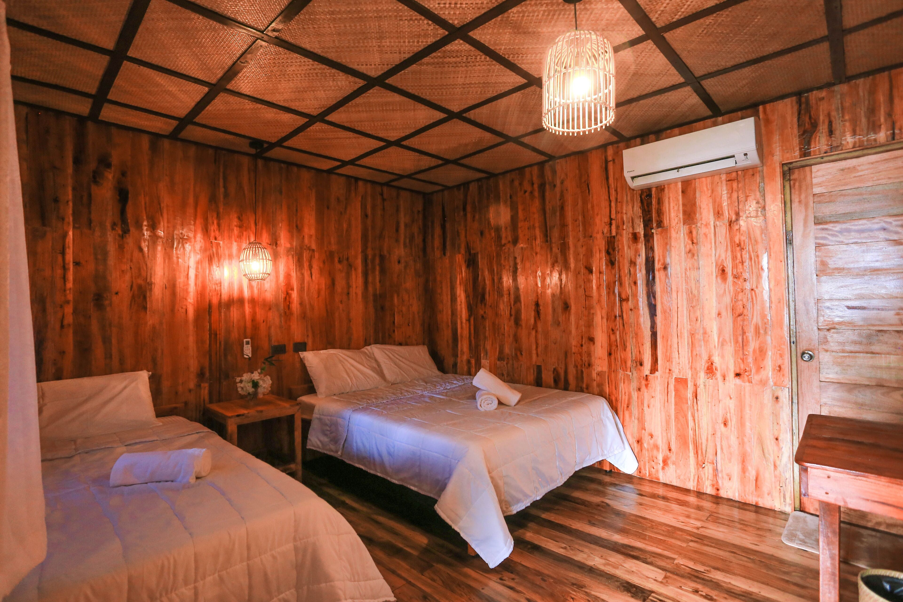 A room with wooden walls and a ceiling with a grid pattern. There are two beds with white bedding and a small table with a lamp. An air conditioning unit is mounted on the wall.