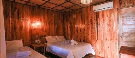 A room with wooden walls and a ceiling with a grid pattern. There are two beds with white bedding and a small table with a lamp. An air conditioning unit is mounted on the wall.